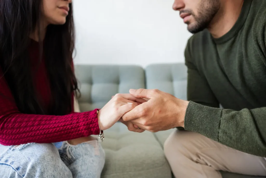 A couple demonstrating emotional intelligence through physical connection, holding hands intimately while engaged in conversation on a light gray couch. The person in the burgundy sweater and the person in the olive green sweater share a moment of empathy and understanding, illustrating how emotional intelligence enables meaningful relationships and vulnerable communication.