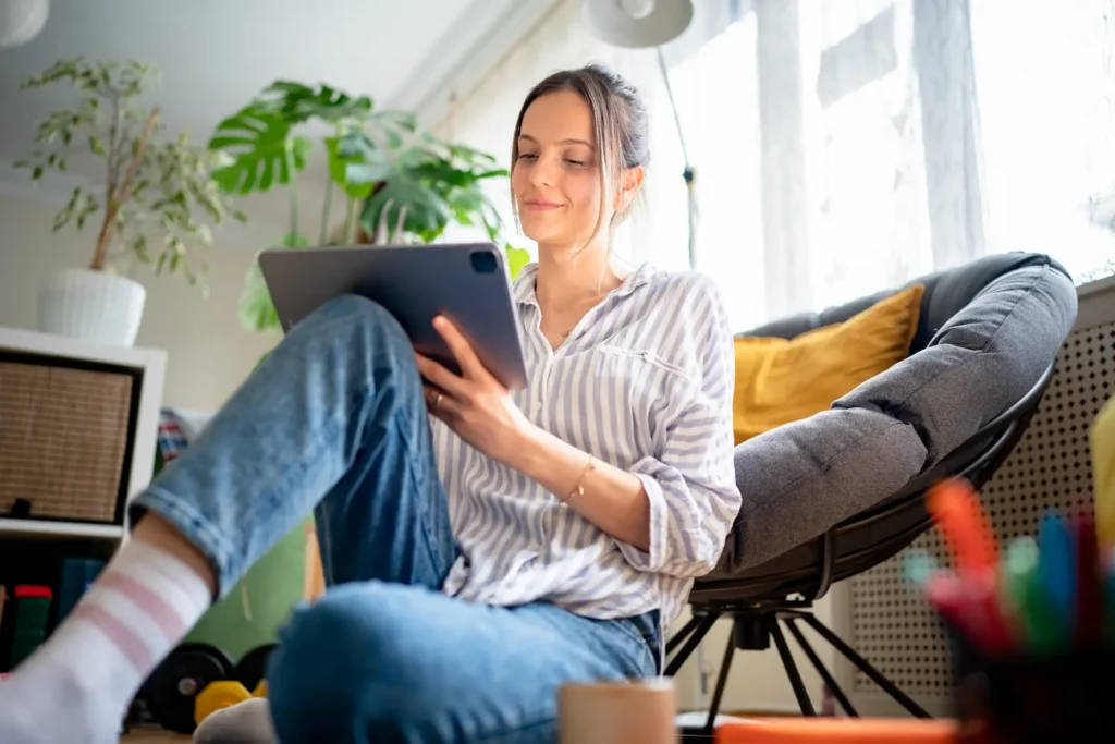 Young woman smiling while browsing on a tablet at home, relaxing in a comfortable chair surrounded by plants and natural light – representing the fulfilling solo lifestyle that challenges the 'why are you still single' question while showcasing independence and contentment in her own space.